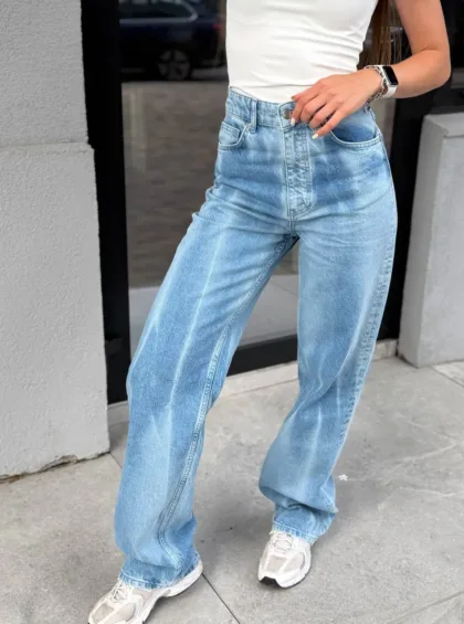 Person wearing light-wash high-waisted jeans with a white top, standing on a sidewalk outdoors.