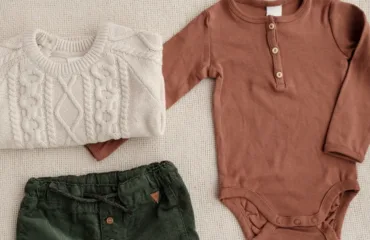 Yak vybraty dytyachyy odyah do sadochka abo shkoly komfort rozmir tkanyny Baby clothes laid out on a beige surface: cream knitted sweater, rust long-sleeve onesie, and dark green drawstring pants.