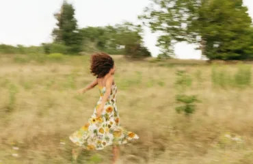 Person in a floral sundress running through a sunlit field, hair in motion as the grass sways around her/they stay blurred by movement.