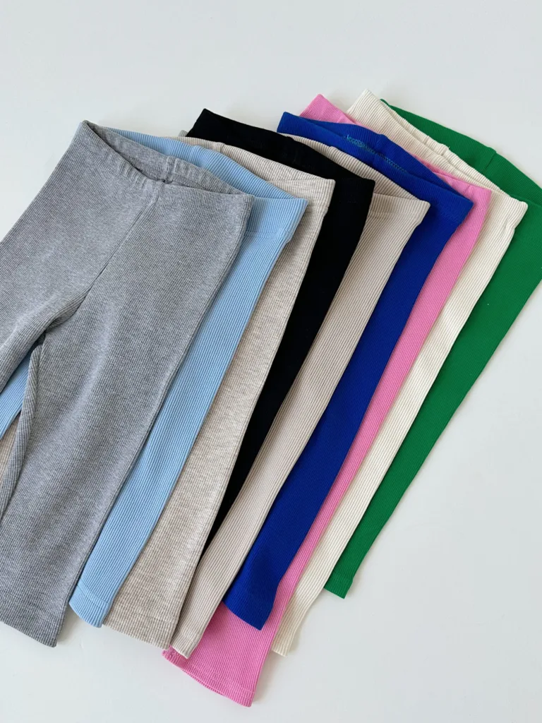 Одяг для дітей до садочка та школи: як обрати - Row of folded fabric swatches in gray, light blue, beige, black, cream, blue, pink, white, and green arranged in a curved line on a light background.