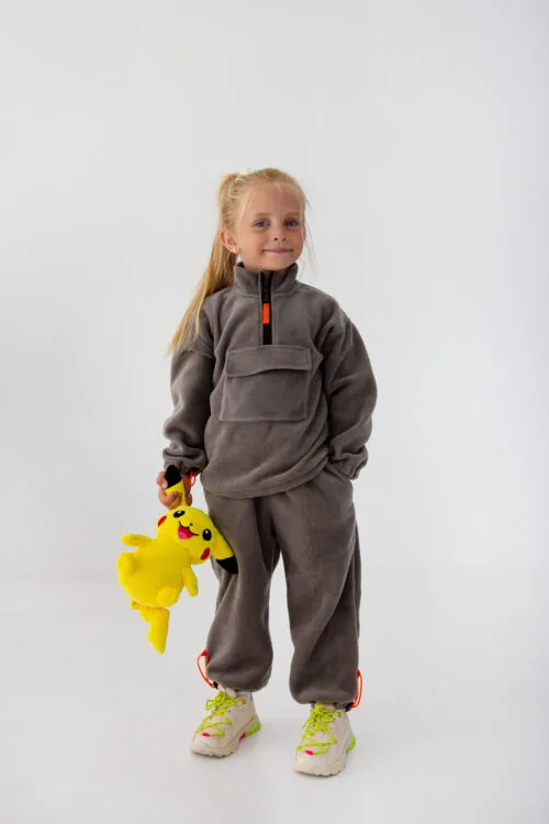 Одяг для дітей до садочка та школи: як обрати - Young girl in a gray fleece jumpsuit holding a Pikachu plush toy against a white background.