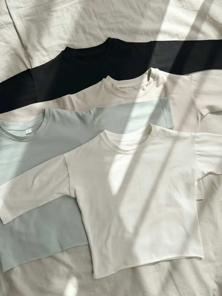 Одяг для дітей до садочка та школи: як обрати - Four long-sleeve crewneck shirts in black, beige, pale blue, and white laid diagonally on a bed.