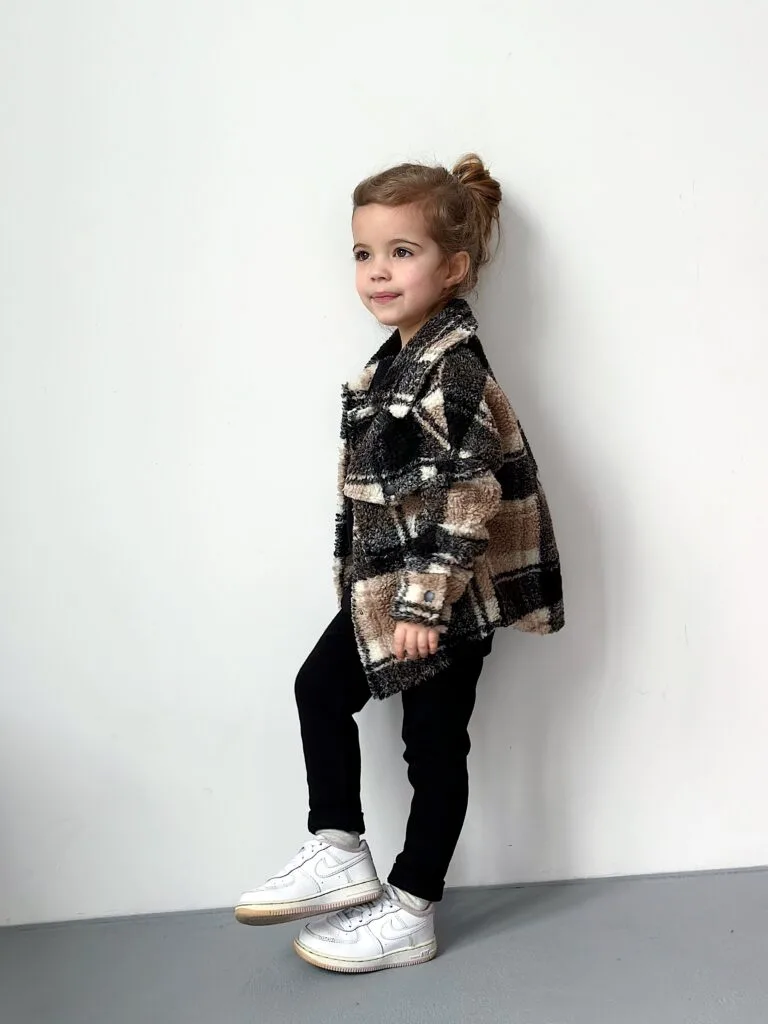 Одяг для дітей до садочка та школи: як обрати - Young girl wearing a black-and-beige plaid coat, black pants, and white sneakers standing against a white wall.