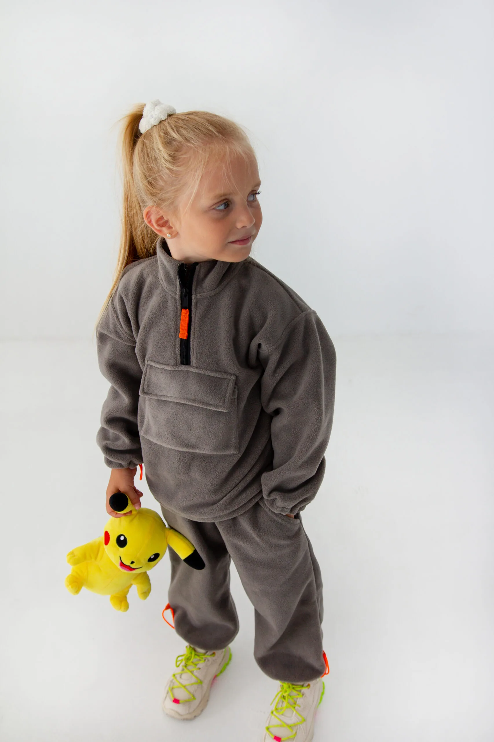 Одяг для дітей до садочка та школи: як обрати - Young girl in a gray fleece jumpsuit looks to the right, holding a yellow Pikachu plush toy