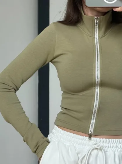 Cropped olive-green zip-up long-sleeve top with a silver zipper, paired with white drawstring shorts in a fashion pose.