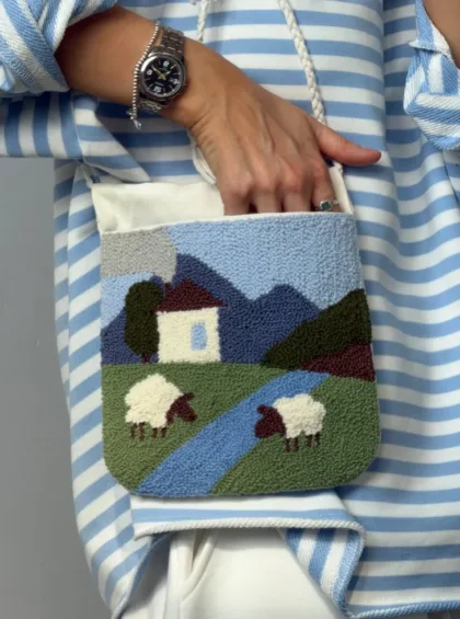 photo_5400255715378667294_y Person wearing a blue-and-white striped shirt reaches into a small tapestry bag featuring a countryside scene with a house, a river, hills, and two sheep on grass.