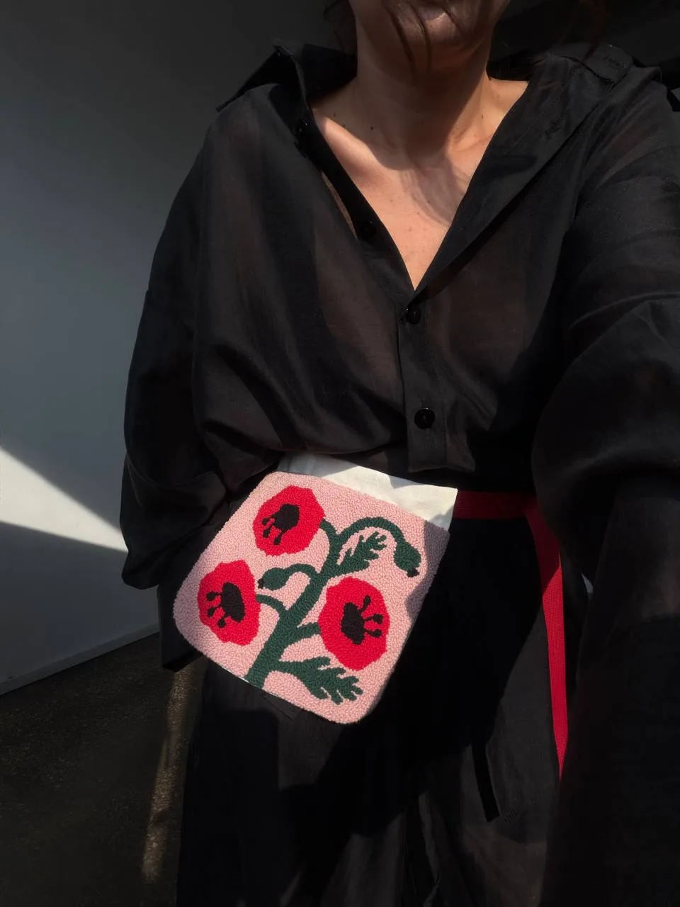 Кишенька «Маки» за ціною 1990.00 ₴ в магазині Romanova Shop.Сучасний погляд на традиційний аксесуар Вашій увазі — ладунка! Або гаман, кишеня чи кишенька — назва залежить від регіону, але суть одна: невелика торбинка для найважливішого. Колись у них носили монети, ключі, обереги та пам’ятні дрібнички. Сьогодні ми переосмислили цей традиційний аксесуар і створили колекцію ладунок із килимовою вишивкою — для телефону, карток, навушників, помади чи всіх тих речей, які хочеться тримати під рукою. Всередині на вас чекає маленький сюрприз: кожна ладунка має лляну підкладку свого кольору. Яка саме дістанеться вам — заздалегідь невідомо. Несподівана деталь, яка робить кожну річ ще більш особливою. У комплекті ви знайдете плетений бавовняний шнурок та контрастну ремінну стрічку, щоб обрати спосіб носіння, який пасує саме вам. - photo_5400255715378667315_y Купити Кишенька «Маки» - Romanova - Person in a black button-down shirt holding a pink square floral pouch at the waist featuring red flowers and green stems.