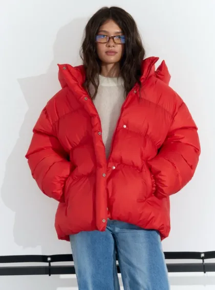 Person wearing a bright red oversized puffer jacket with hands in pockets, standing against a white wall.