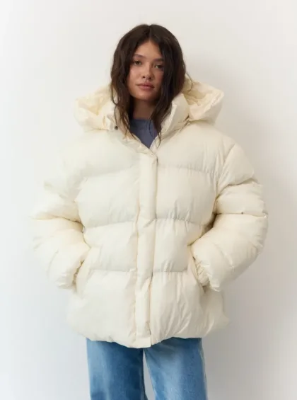 Woman wearing an oversized cream puffer jacket with hood, hands in pockets, standing against a white wall.