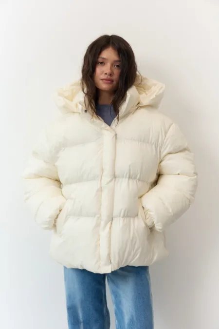 Пуховик чорний жіночий оверсайз екопух до -15°C XS/S M/L - Woman wearing an oversized cream puffer jacket with hood, hands in pockets, standing against a white wall.