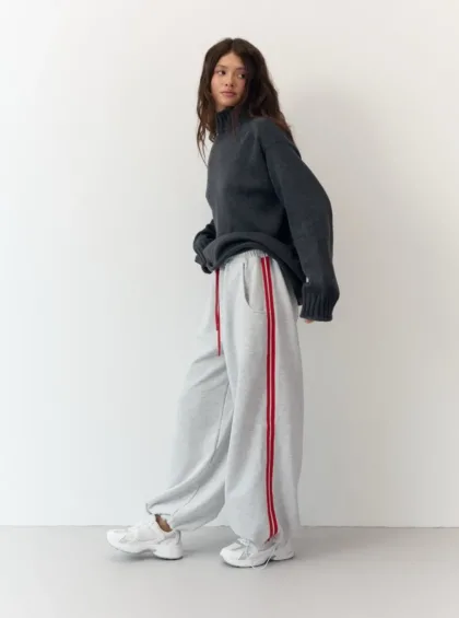 Woman wearing a dark gray oversized sweater and light gray joggers with red side stripes, standing against a white wall.