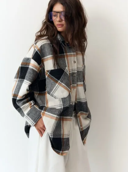 Casual fashion shot: a model wearing an oversized plaid flannel shirt in neutral tones with white pants and blue-tinted sunglasses, posing against a white background.