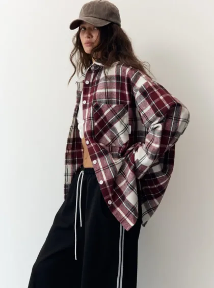 Person in an oversized burgundy plaid shirt over a white top, black drawstring pants, and a brown cap, posing against a plain wall.