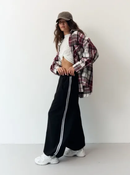 Casual streetwear: cap, burgundy plaid shirt over a knotted white tee, black wide-leg pants with white stripes, and chunky sneakers.