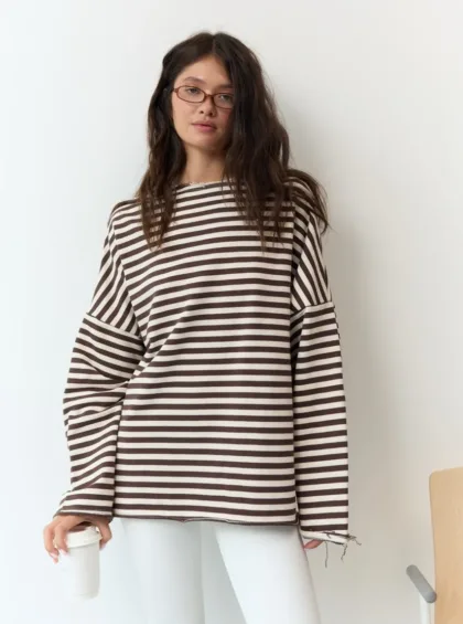 Young woman with brown hair and glasses wearing a brown-and-white striped oversized sweater, holding a white coffee cup against a white wall.