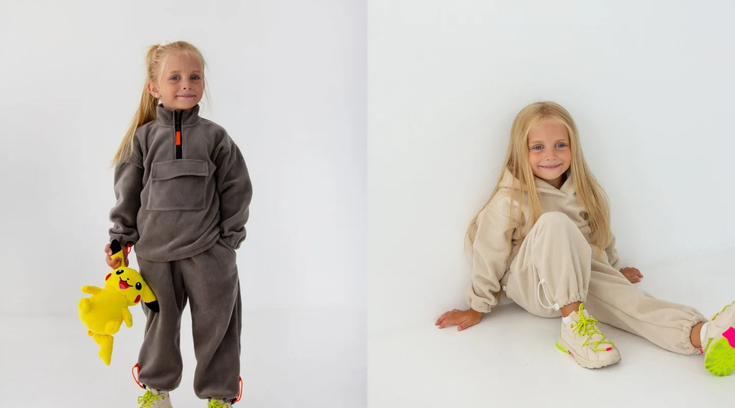 Dytyachi kostyumy na osin yak pidibraty komplekt dlya shkoly sadochka y prohulyanok Two young girls in neutral loungewear pose against a white backdrop; one stands holding a yellow Pikachu plush toy, the other sits on the floor.