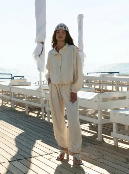 Woman standing on a sunlit seaside boardwalk in a cream tracksuit and grid headpiece, with the ocean in the background.