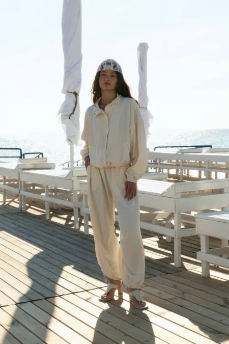 Костюм жіночий поло молочний oversize з вишивкою - Woman standing on a sunlit seaside boardwalk in a cream tracksuit and grid headpiece, with the ocean in the background.