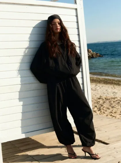 Person wearing all-black outfit and headscarf leaning against a white beach shack by the sea, wearing black loose pants and flip-flops.