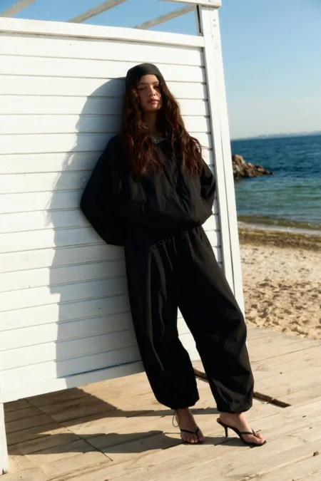 Костюм жіночий графітовий oversize з футеру XS/S M/L - Person wearing all-black outfit and headscarf leaning against a white beach shack by the sea, wearing black loose pants and flip-flops.