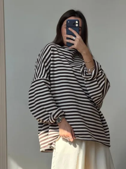 Person taking a mirror selfie in an oversized black-and-white striped sweater with a light skirt, wearing silver jewelry (watch and bracelets) and holding a dark blue phone.