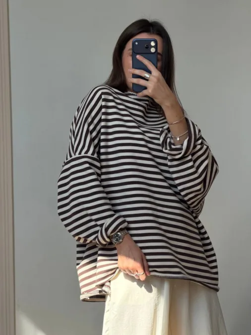 Лонг жіночий молочний з принтом Rebelde way бавовна - Person taking a mirror selfie in an oversized black-and-white striped sweater with a light skirt, wearing silver jewelry (watch and bracelets) and holding a dark blue phone.