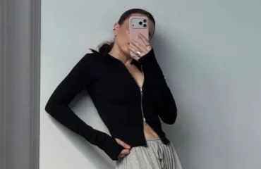 Person taking a mirror selfie wearing a black zip-up top and gray sweatpants, hand on hip, phone covering face.