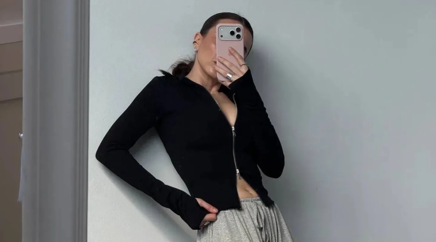 Lonhsliv zhinochyy chomu tse nayzruchnisha rich dlya mizhsezonnya Person taking a mirror selfie wearing a black zip-up top and gray sweatpants, hand on hip, phone covering face.