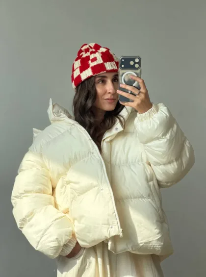 Woman taking a mirror selfie in a cream puffer jacket and a red-and-white checkered knit beanie.