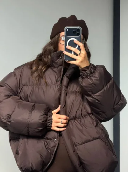 Person taking a mirror selfie in a brown oversized puffer jacket and matching brown beret, obscuring their face with a smartphone case with a white circular logo on it.