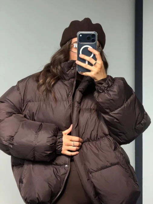 Бомбер жіночий флісовий червоний one size полар-фліс - Person taking a mirror selfie in a brown oversized puffer jacket and matching brown beret, obscuring their face with a smartphone case with a white circular logo on it.