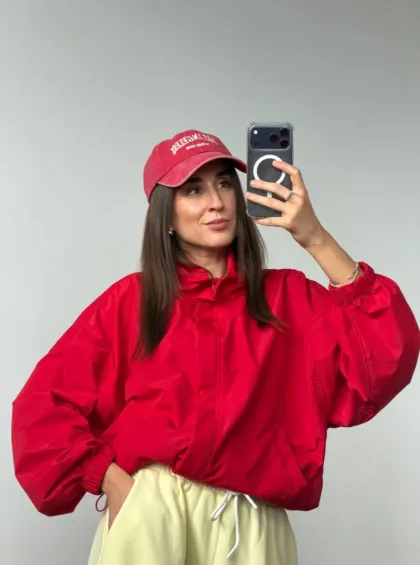 Woman in a red windbreaker and matching cap taking a mirror selfie with a smartphone against a gray wall.