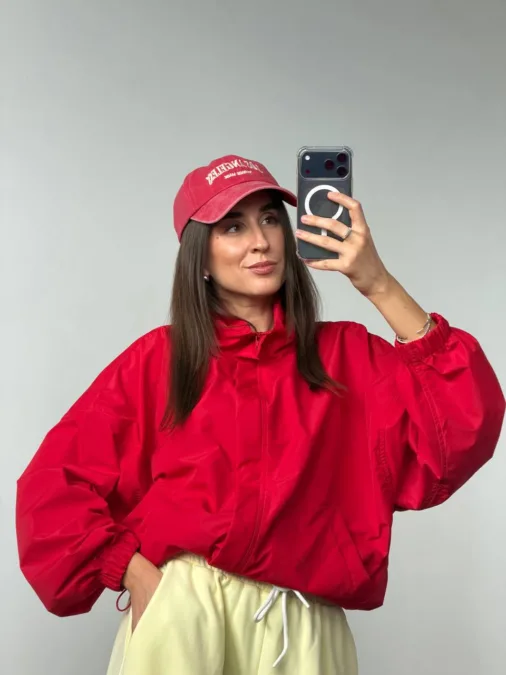 Бомбер жіночий флісовий оливковий one size полар-фліс - Woman in a red windbreaker and matching cap taking a mirror selfie with a smartphone against a gray wall.