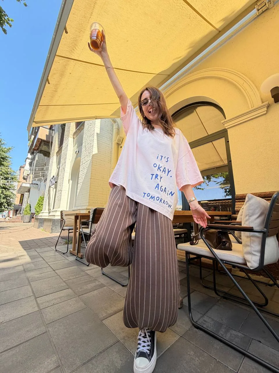 Ніжно-рожева футболка з принтом It`s okay. Try Again tomorrow від Romanova - бавовна 95%, еластан 5%, розмір one size. Довжина 69 см, ширина 64 см. - photo_5433703400261491097_y Футболка жіноча ніжно-рожева з принтом It`s okay бавовна - Young woman in a white shirt with the text 'It’s OKAY, TRY AGAIN TOMORROW' raises a bottle over her head at a sunny sidewalk café.
