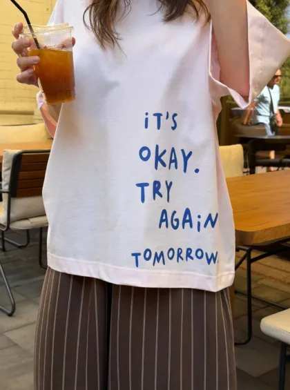 photo_5433703400261491099_y Person at an outdoor cafe wears a white shirt with blue text reading 'It's okay. Try again tomorrow' while holding an iced coffee with a straw.