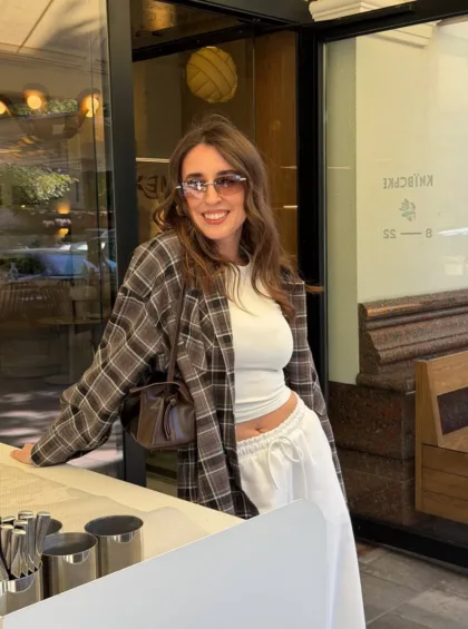 Smiling woman in sunglasses leaning on a cafe counter, wearing a white crop top, white drawstring pants, and a plaid shirt with a brown handbag.