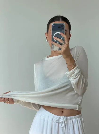 Person in a white cropped long-sleeve top and white drawstring shorts taking a mirror selfie with a phone, standing against a light wall.