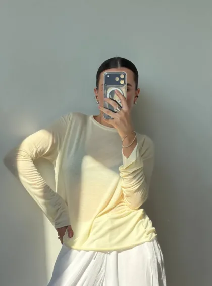 Person posing for a mirror selfie in a pale yellow long-sleeve top and white skirt, holding a phone to cover their face.