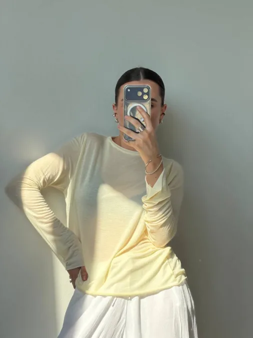 Лонгслів жіночий молочний приталений напівпрозорий XS/S M/L - Person posing for a mirror selfie in a pale yellow long-sleeve top and white skirt, holding a phone to cover their face.