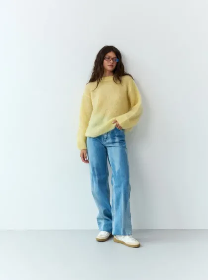 Woman posing against a plain white wall in a pale yellow fuzzy sweater, blue jeans, and white sneakers, with glasses.