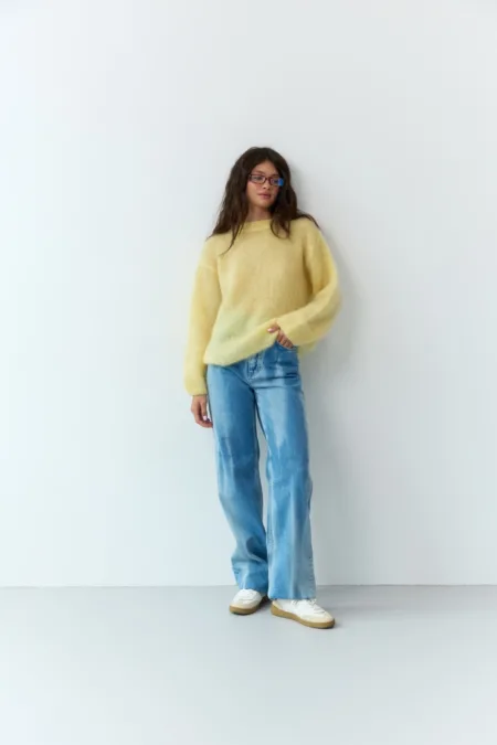 Жіночий джинс светр зі 95% merino kids, розмір one size. Мʼякий теплий трикотаж для базових повсякденних образів. Виробництво Україна. - 1C7A7651 велике Светр жіночий джинс 95% merino kids one size - Woman posing against a plain white wall in a pale yellow fuzzy sweater, blue jeans, and white sneakers, with glasses.