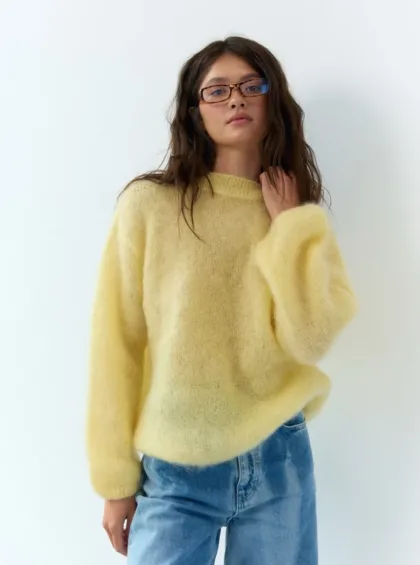 Woman wearing a pale yellow fuzzy sweater and blue jeans, posing against a white wall.