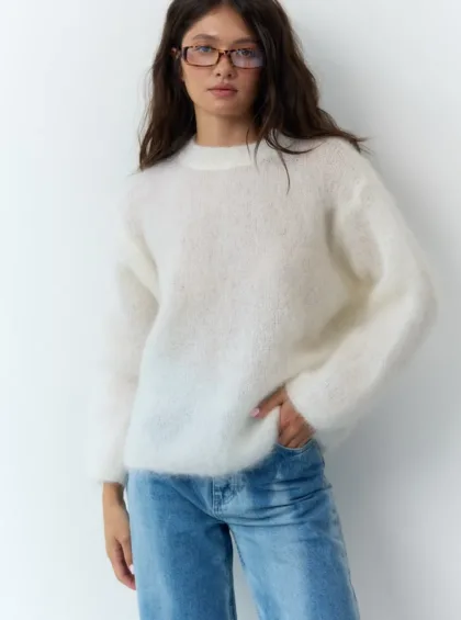 Woman with dark hair and tortoiseshell glasses wearing a fluffy white knit sweater and light blue jeans, standing against a light wall.