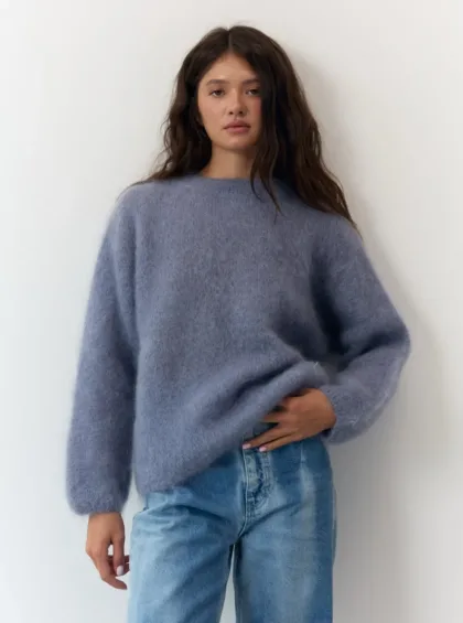 Woman wearing an oversized fuzzy blue sweater and light-wash jeans, standing against a white wall and looking at the camera.