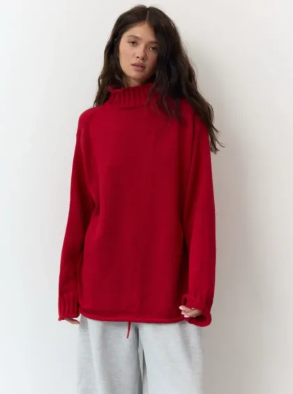 Woman wearing an oversized red turtleneck sweater and light gray sweatpants, standing against a white wall.