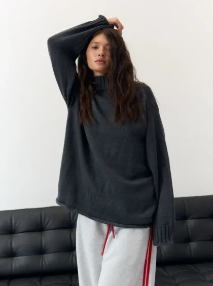 Woman in oversized dark gray sweater and light gray joggers with red drawstrings, posing with one arm raised by a white wall.