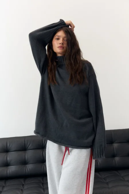 Светр жіночий оливковий бавовняний one size - Woman in oversized dark gray sweater and light gray joggers with red drawstrings, posing with one arm raised by a white wall.