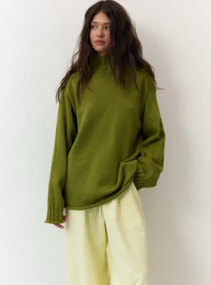 Woman wearing an olive knit sweater and light cream pants, standing against a white wall with hands in pockets/light pose.