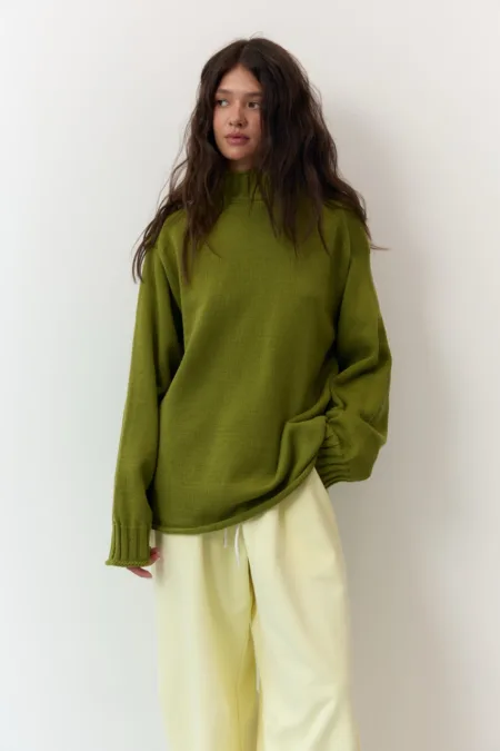 Укорочене пальто сіре oversize із вовни - Woman wearing an olive knit sweater and light cream pants, standing against a white wall with hands in pockets/light pose.