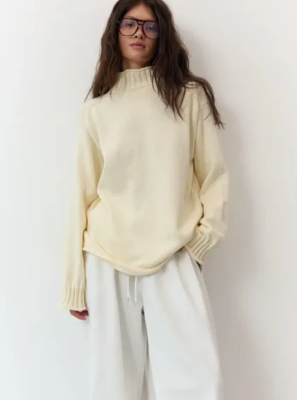 Woman modeling a cream knit sweater and white wide-leg pants, wearing glasses, standing against a light background.